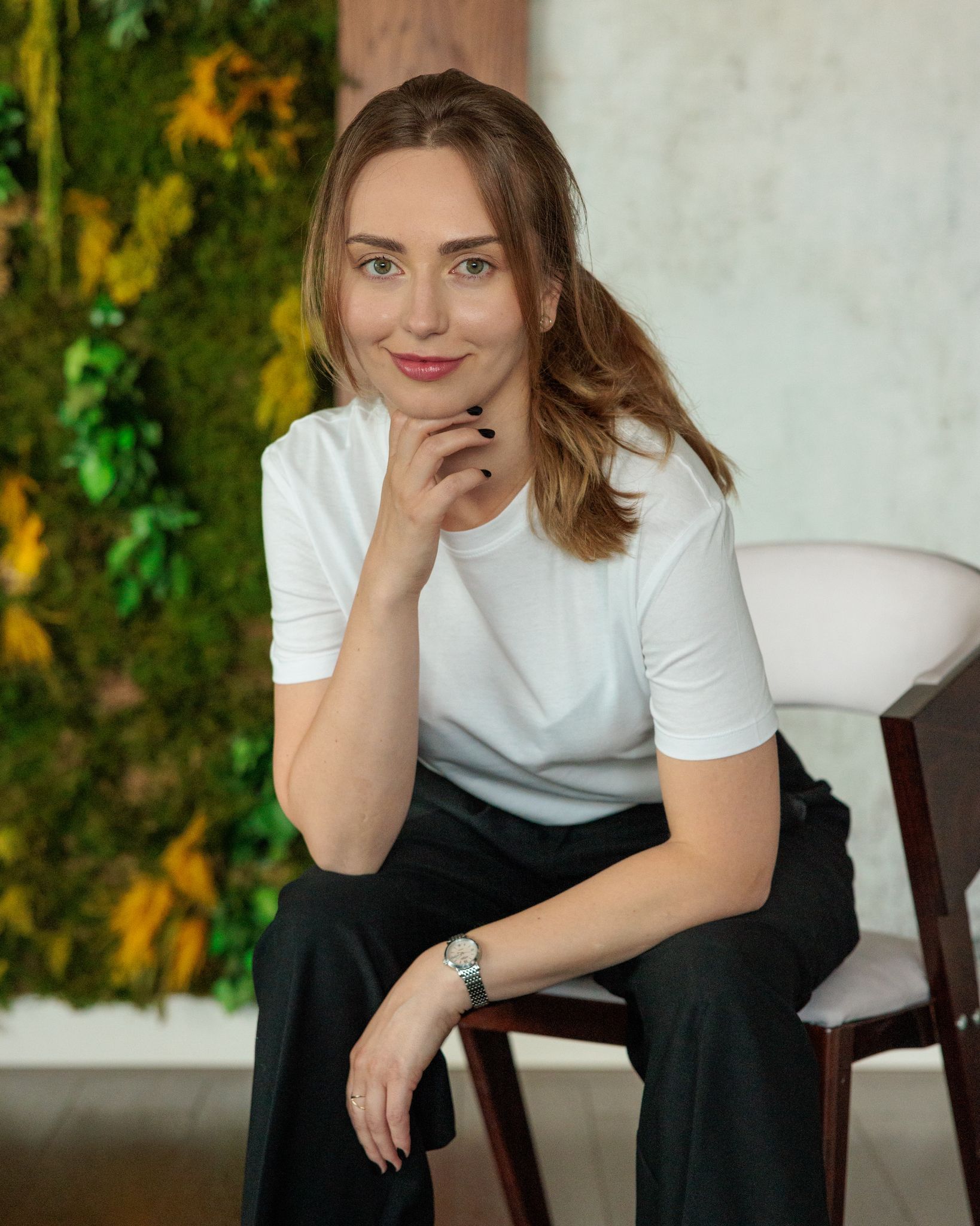 Anastasia Goluk siedzi na krześle na tle zielonej ściany z mchu, uśmiecha się ciepło